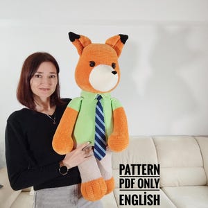 May include: A large, orange and white crocheted fox doll wearing a green shirt and a blue striped tie. The fox has black-tipped ears and is holding a pattern. The text "PATTERN PDF ONLY ENGLISH" is visible.