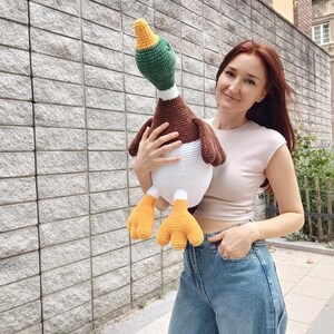 May include: A large, handmade crochet duck with a green head, brown body, white belly, and orange feet. The duck is being held in front of a gray stone wall. The duck's head is turned slightly to the side.
