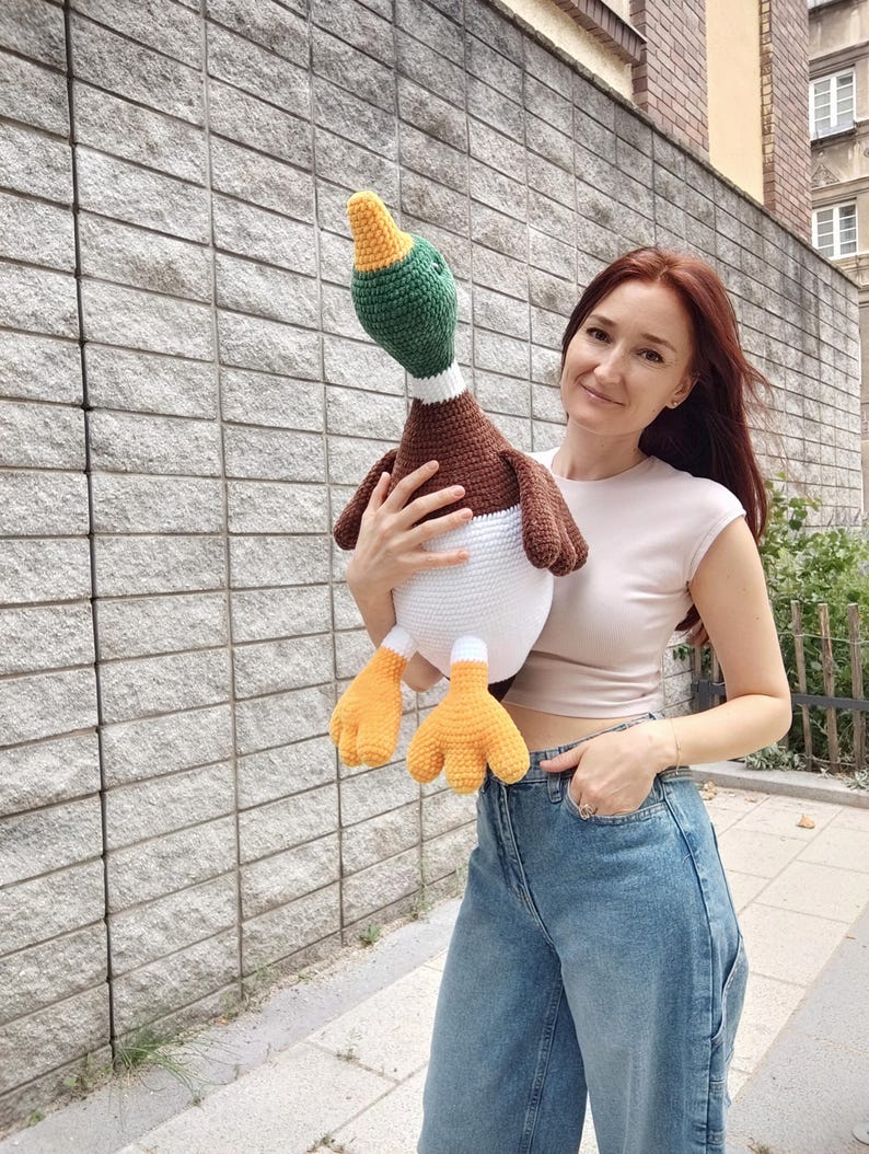 May include: A large, handmade crochet duck with a green head, brown body, white belly, and orange feet. The duck is being held in front of a gray stone wall. The duck's head is turned slightly to the side.