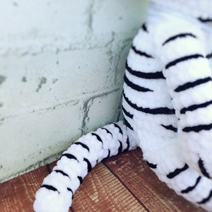 May include: A white and black striped plush toy tiger, with a focus on the tail and leg. The toy is made of a soft, textured material. The background is a light-colored brick wall and a wooden surface.