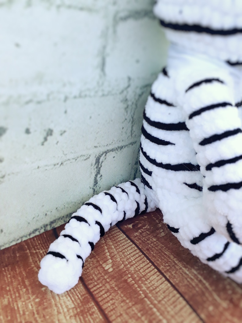 May include: A white and black striped plush toy tiger, with a focus on the tail and leg. The toy is made of a soft, textured material. The background is a light-colored brick wall and a wooden surface.