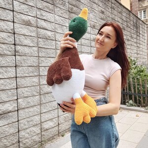 May include: A large, handmade crochet duck toy. The duck features a green head with a yellow beak, brown wings, a white body, and yellow feet. The toy is held up against a gray brick wall.