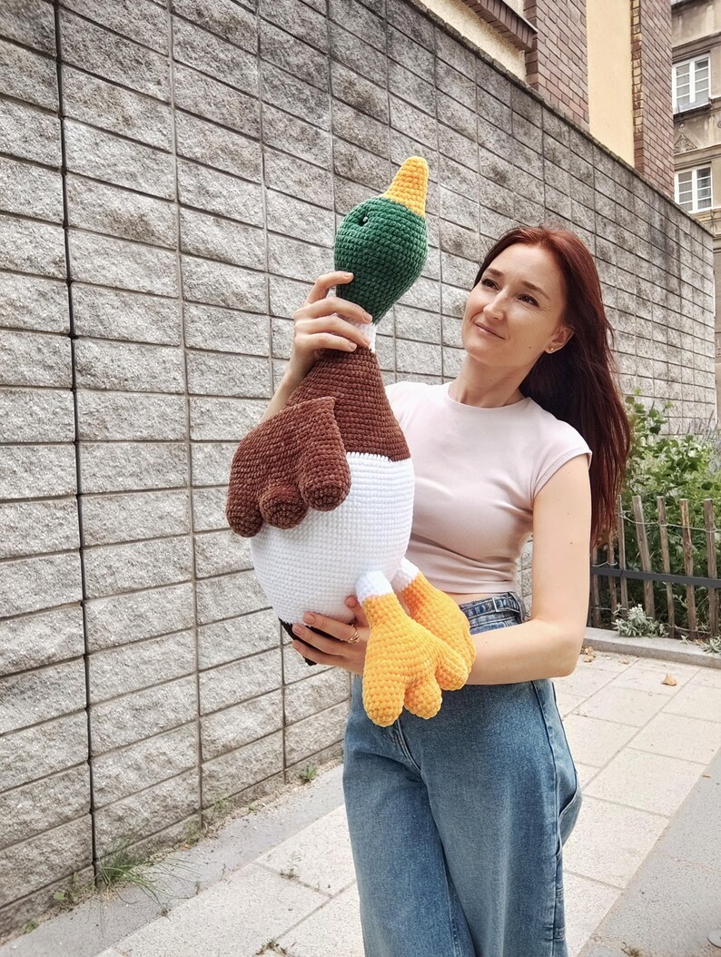 May include: A large, handmade crochet duck toy. The duck features a green head with a yellow beak, brown wings, a white body, and yellow feet. The toy is held up against a gray brick wall.