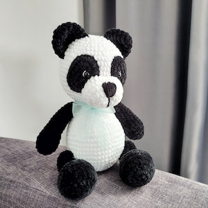 K&ouml;nnte beinhalten: Ein handgefertigter Panda-B&auml;r aus Pl&uuml;sch mit wei&szlig;em K&ouml;rper, schwarzen Akzenten und einem hellblauen Band. Der Panda hat schwarze Ohren, Augen und Gliedma&szlig;en. Das Stofftier sitzt auf einer grauen Oberfl&auml;che.