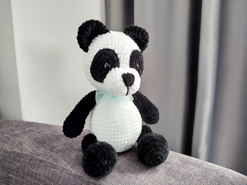 K&ouml;nnte beinhalten: Ein handgefertigter Panda-B&auml;r aus Pl&uuml;sch mit wei&szlig;em K&ouml;rper, schwarzen Akzenten und einem hellblauen Band. Der Panda hat schwarze Ohren, Augen und Gliedma&szlig;en. Das Stofftier sitzt auf einer grauen Oberfl&auml;che.