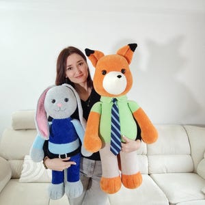 May include: Two large, handmade crochet toys: a grey bunny with pink ears and a blue outfit, and an orange fox wearing a green shirt and blue striped tie. The fox has black accents on its ears. Both are held by a person.