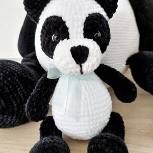 K&ouml;nnte beinhalten: Ein handgefertigtes Panda-B&auml;r-Pl&uuml;schtier mit schwarz-wei&szlig;em Fell. Der Panda hat schwarze Ohren, Augen und Gliedma&szlig;en, mit einem wei&szlig;en K&ouml;rper und Kopf. Ein hellblaues Band ist um seinen Hals gebunden. Der Panda sitzt auf einer hellen Holzoberfl&auml;che.