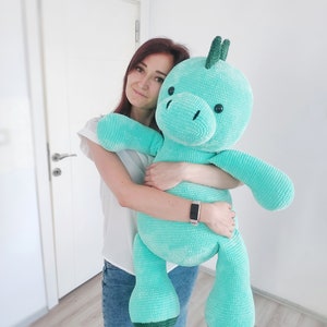 May include: A large, turquoise plush toy in the shape of a friendly dinosaur. The stuffed animal has a textured, knitted appearance, with dark green accents on the horns and feet. The toy is being held in front of a white door.