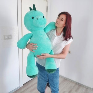 May include: A large, turquoise crocheted dinosaur plush toy with a green spine and feet is held by a person. The dinosaur has a round head, small black eyes, and a long neck. The plush toy is a soft, cuddly design.
