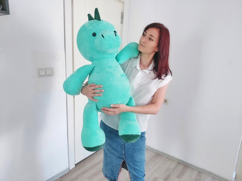 May include: A large, turquoise crocheted dinosaur plush toy with a green spine and feet is held by a person. The dinosaur has a round head, small black eyes, and a long neck. The plush toy is a soft, cuddly design.