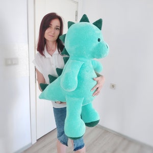 May include: A large, turquoise crocheted dinosaur plush toy with dark green accents. The stuffed animal features a long tail, spiky dorsal fins, and a friendly expression. The toy is approximately 36 inches tall.