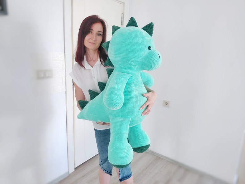 May include: A large, turquoise crocheted dinosaur plush toy with dark green accents. The stuffed animal features a long tail, spiky dorsal fins, and a friendly expression. The toy is approximately 36 inches tall.