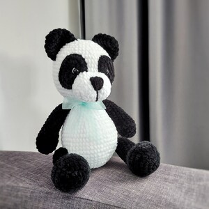 K&ouml;nnte beinhalten: Ein handgefertigtes Panda-B&auml;r-Pl&uuml;schtier aus H&auml;kelarbeit. Der Panda hat einen wei&szlig;en K&ouml;rper, schwarze Arme, Beine und Ohren sowie schwarze Kreise um die Augen. Er hat ein kleines, hellblaues Band um den Hals gebunden. Das Pl&uuml;schtier sitzt auf einer grauen Oberfl&auml;che.