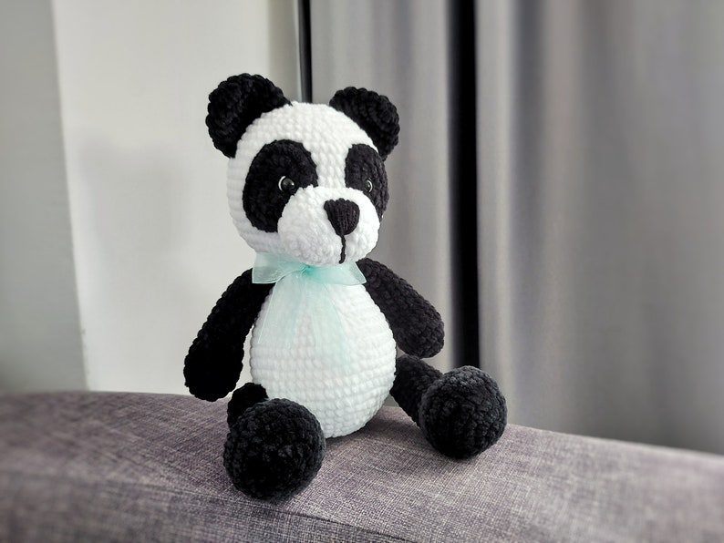 K&ouml;nnte beinhalten: Ein handgefertigtes Panda-B&auml;r-Pl&uuml;schtier aus H&auml;kelarbeit. Der Panda hat einen wei&szlig;en K&ouml;rper, schwarze Arme, Beine und Ohren sowie schwarze Kreise um die Augen. Er hat ein kleines, hellblaues Band um den Hals gebunden. Das Pl&uuml;schtier sitzt auf einer grauen Oberfl&auml;che.