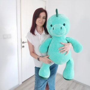 May include: A large, turquoise crocheted dinosaur plush toy with a rounded head, small black eyes, and a green spine. The plush toy is being held by a person. The toy has a soft, textured appearance, and is a handmade item.