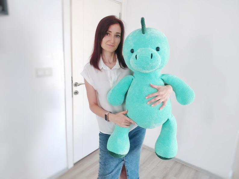 May include: A large, turquoise crocheted dinosaur plush toy with a rounded head, small black eyes, and a green spine. The plush toy is being held by a person. The toy has a soft, textured appearance, and is a handmade item.