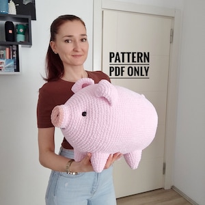 May include: A large, light pink crocheted pig plush toy with black eyes and a pink snout. The pig has small ears and stubby legs. The text "PATTERN PDF ONLY" is visible in the background. This handmade item is a soft, cuddly toy.