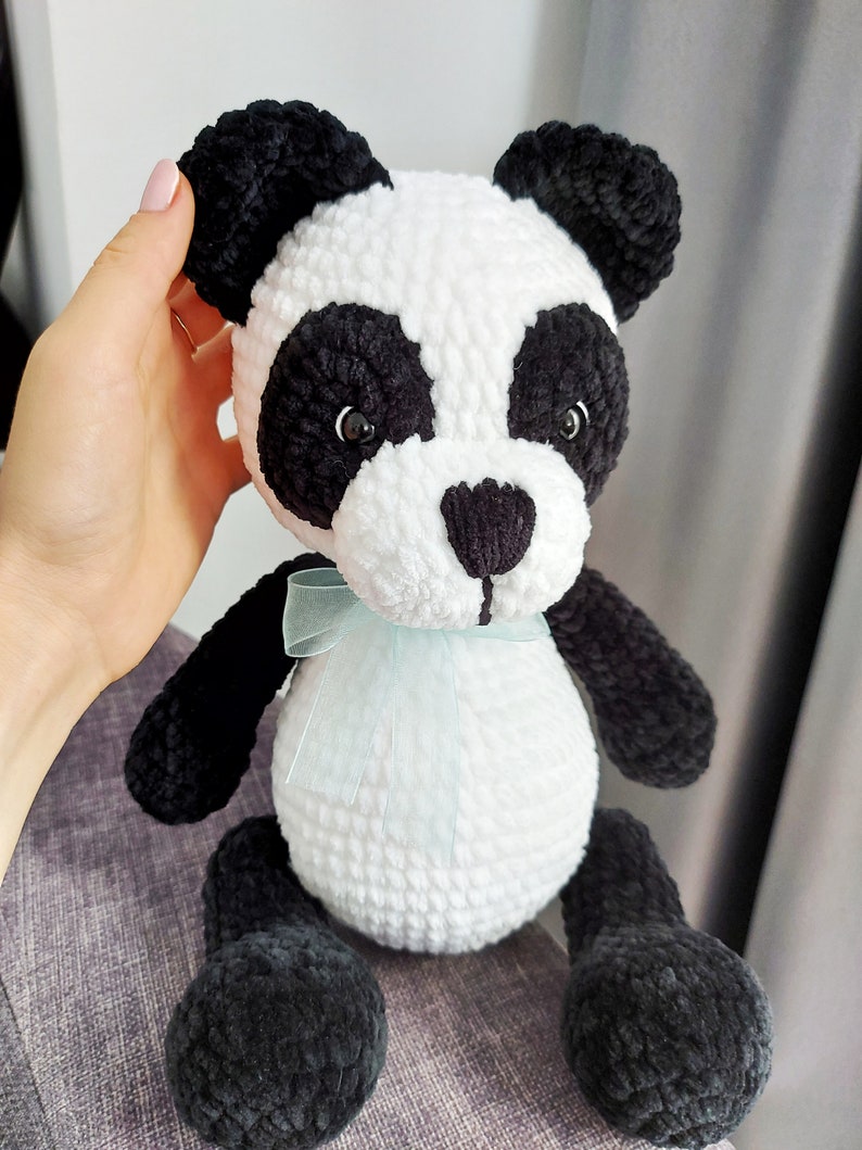 K&ouml;nnte beinhalten: Ein handgefertigter Panda-B&auml;r aus Pl&uuml;sch mit schwarz-wei&szlig;em Fell. Der Panda hat schwarze Ohren, Augen und Gliedma&szlig;en, mit einem wei&szlig;en Gesicht und K&ouml;rper. Eine hellblaue Schleife ist um seinen Hals gebunden. Das Spielzeug ist weich und kuschelig.
