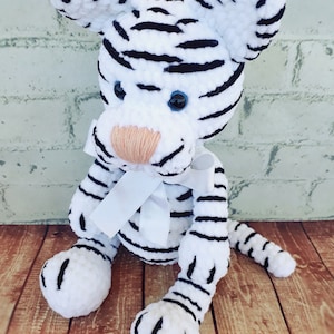 May include: A handmade white tiger plush toy with black stripes, blue eyes, and a peach-colored nose. The tiger has a white bow and is sitting on a wooden surface. The plush toy is a soft, cuddly animal.