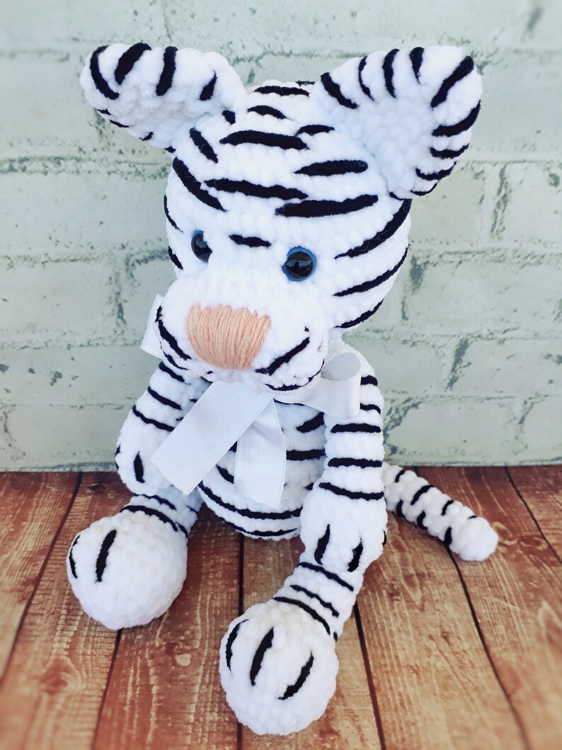 May include: A handmade white tiger plush toy with black stripes, blue eyes, and a peach-colored nose. The tiger has a white bow and is sitting on a wooden surface. The plush toy is a soft, cuddly animal.