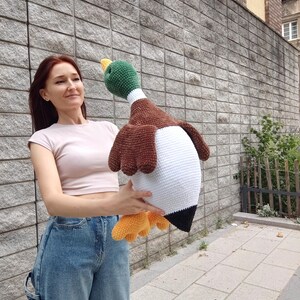 May include: A large, handmade crochet duck with a green head, brown wings, and a white body. The duck has yellow feet and a black tail. The plush toy is held up in front of a gray brick wall.
