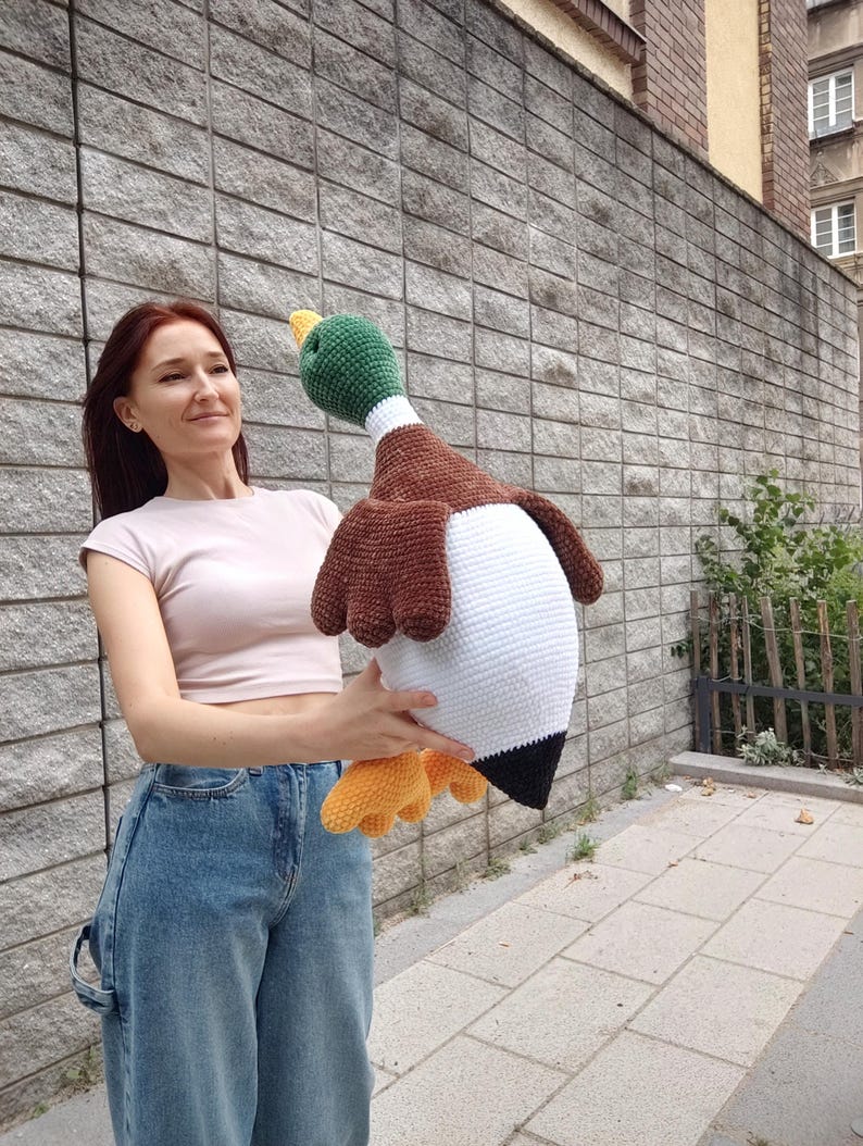 May include: A large, handmade crochet duck with a green head, brown wings, and a white body. The duck has yellow feet and a black tail. The plush toy is held up in front of a gray brick wall.