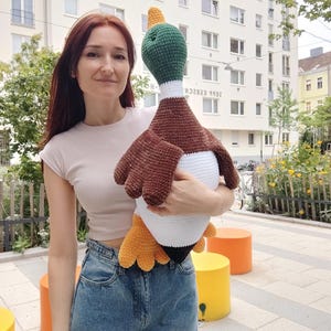 May include: A large, handmade crochet duck plushie. The duck features a green head, brown wings, a white body, and yellow feet. The plushie is held by a person in a light-colored top and blue jeans.