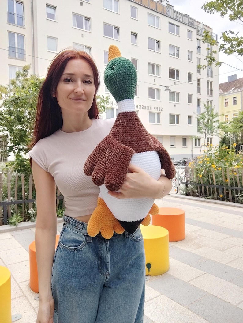 May include: A large, handmade crochet duck plushie. The duck features a green head, brown wings, a white body, and yellow feet. The plushie is held by a person in a light-colored top and blue jeans.