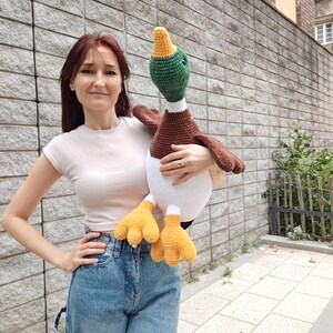 May include: A large, handmade crochet duck plushie. The duck features a white body, brown wings, a green head, and yellow feet. The duck is being held in front of a gray brick wall.