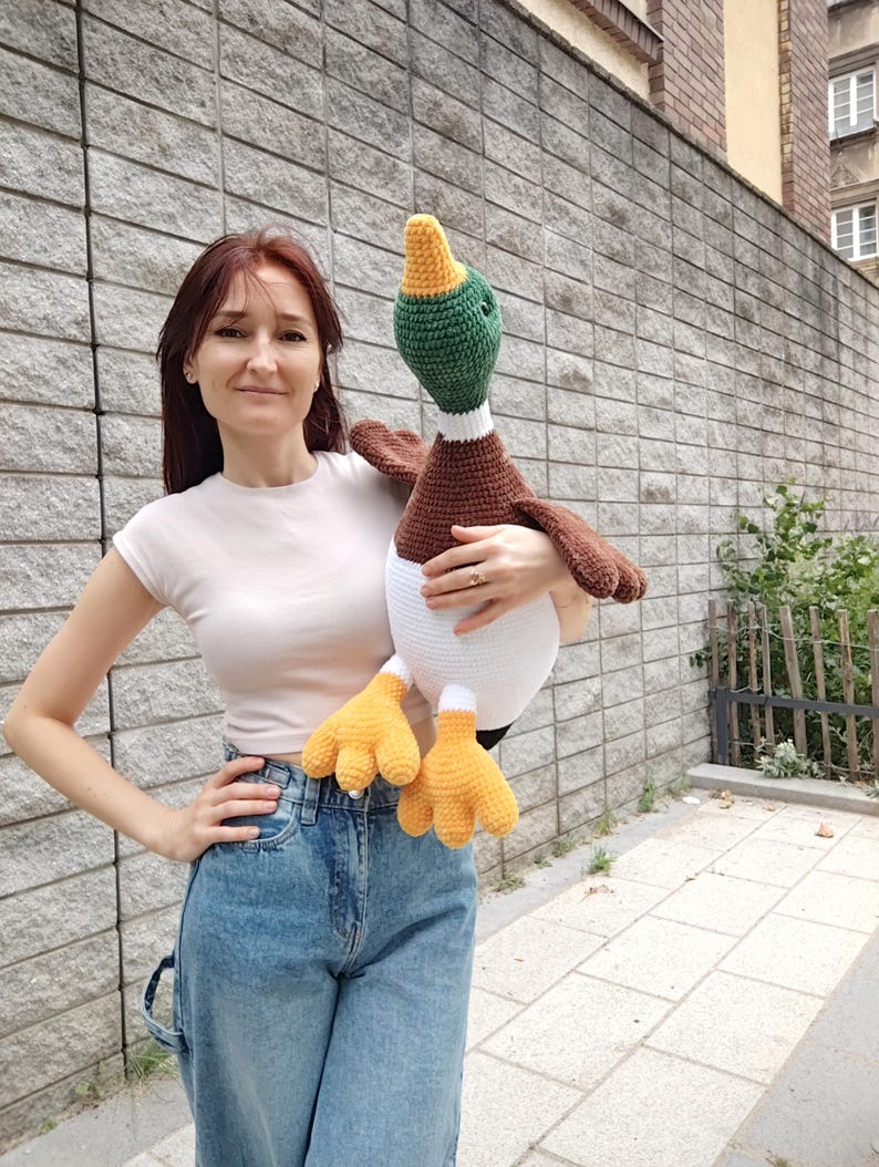 May include: A large, handmade crochet duck plushie. The duck features a white body, brown wings, a green head, and yellow feet. The duck is being held in front of a gray brick wall.