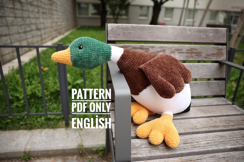 May include: A handmade plush duck with a green head, brown body, and yellow feet. The duck is sitting on a wooden bench. The text "PATTERN PDF ONLY ENGLISH" is visible.
