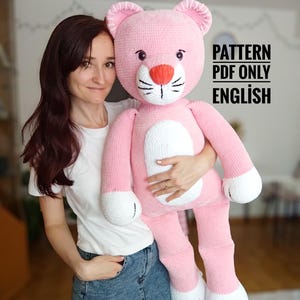 Puede incluir: Un gran oso de peluche rosa hecho a mano con detalles blancos en las patas y el vientre. El oso tiene una nariz roja, bigotes negros y está siendo sostenido. El texto a la derecha dice "PATTERN PDF ONLY ENGLISH".