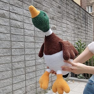 May include: A large, handmade crochet duck plushie. The duck features a green head, brown body, white belly, and orange feet. The duck is being held up in front of a gray brick wall. The duck's height is approximately 18 inches.