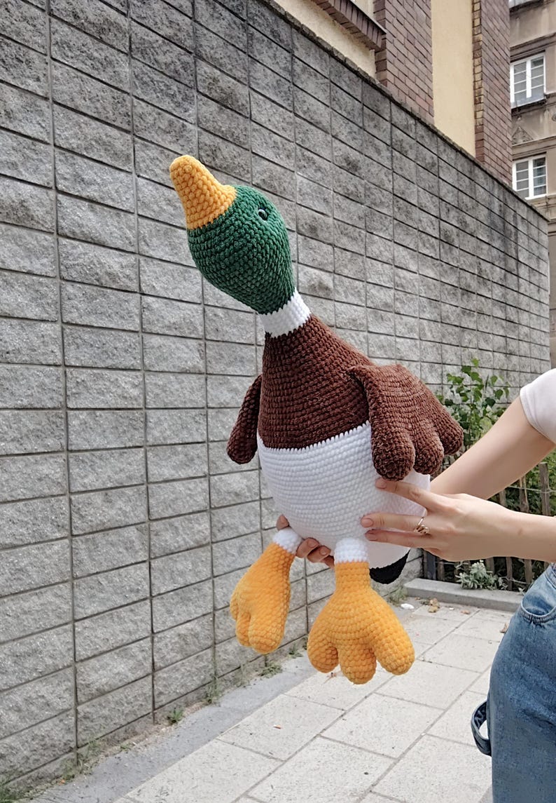 May include: A large, handmade crochet duck plushie. The duck features a green head, brown body, white belly, and orange feet. The duck is being held up in front of a gray brick wall. The duck's height is approximately 18 inches.