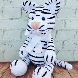 May include: A handmade white and black striped tiger plush toy with a white bow. The tiger has blue eyes and a peach-colored nose. The plush is sitting on a wooden surface.