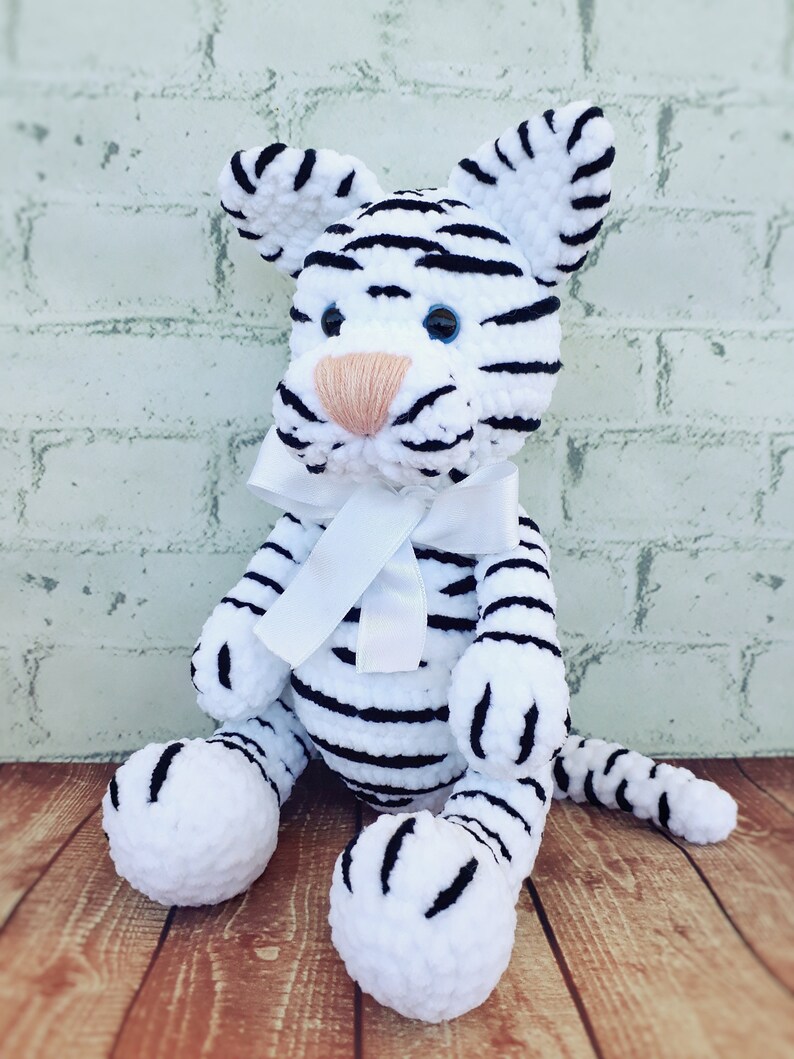 May include: A handmade white and black striped tiger plush toy with a white bow. The tiger has blue eyes and a peach-colored nose. The plush is sitting on a wooden surface.