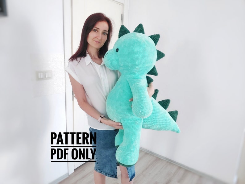May include: A large, turquoise crocheted dinosaur plush toy with dark green spines and accents. The dinosaur is being held by a person. The text "PATTERN PDF ONLY" is visible in the image.