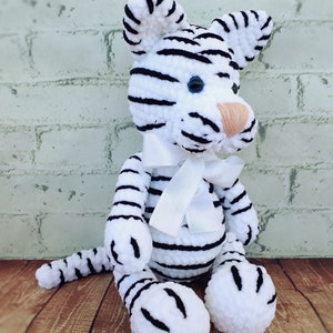 May include: A handmade white tiger plush toy with black stripes, blue eyes, and a peach-colored nose. The tiger has a white ribbon tied around its neck. The plush is sitting on a wooden surface.