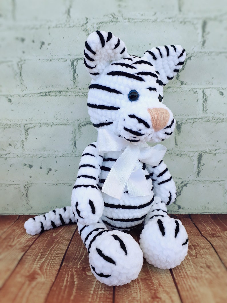 May include: A handmade white tiger plush toy with black stripes, blue eyes, and a peach-colored nose. The tiger has a white ribbon tied around its neck. The plush is sitting on a wooden surface.