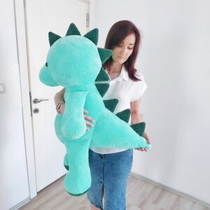 May include: A large, turquoise plush dinosaur with dark green accents. The stuffed animal has a textured, velvety appearance and features a long neck, a rounded head, and a row of triangular spikes down its back. The dinosaur is approximately 36 inches tall.