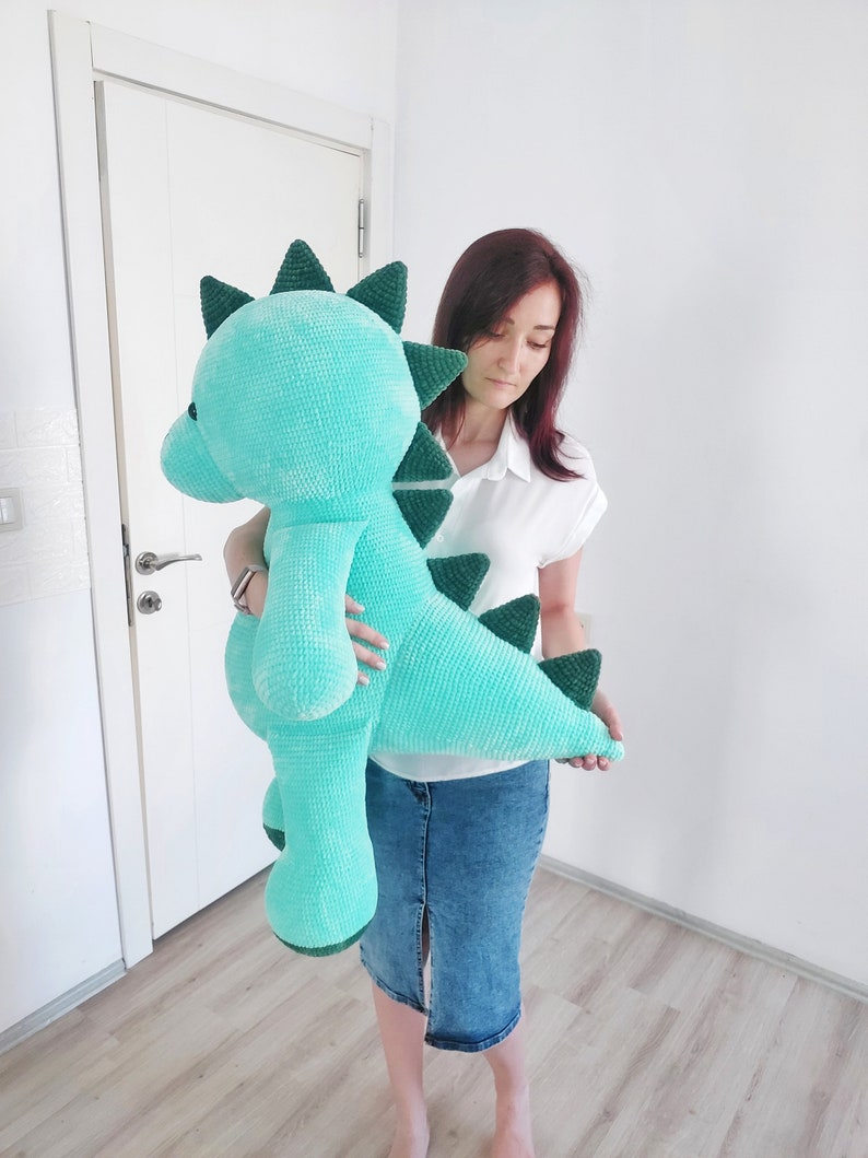 May include: A large, turquoise plush dinosaur with dark green accents. The stuffed animal has a textured, velvety appearance and features a long neck, a rounded head, and a row of triangular spikes down its back. The dinosaur is approximately 36 inches tall.