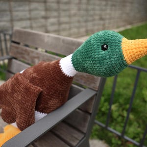 May include: A handmade crochet duck plushie with a green head, brown body, and orange beak. The duck has a white collar and black eyes. The plushie is resting on a wooden bench, showcasing its soft texture and vibrant colors.