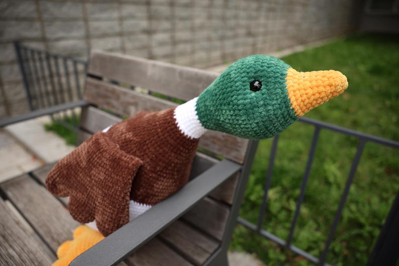 May include: A handmade crochet duck plushie with a green head, brown body, and orange beak. The duck has a white collar and black eyes. The plushie is resting on a wooden bench, showcasing its soft texture and vibrant colors.