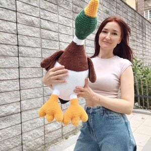 May include: A large, handmade crochet duck plushie. The duck features a brown body, white chest, green head, and yellow beak and feet. The plush toy is held in front of a gray stone wall.