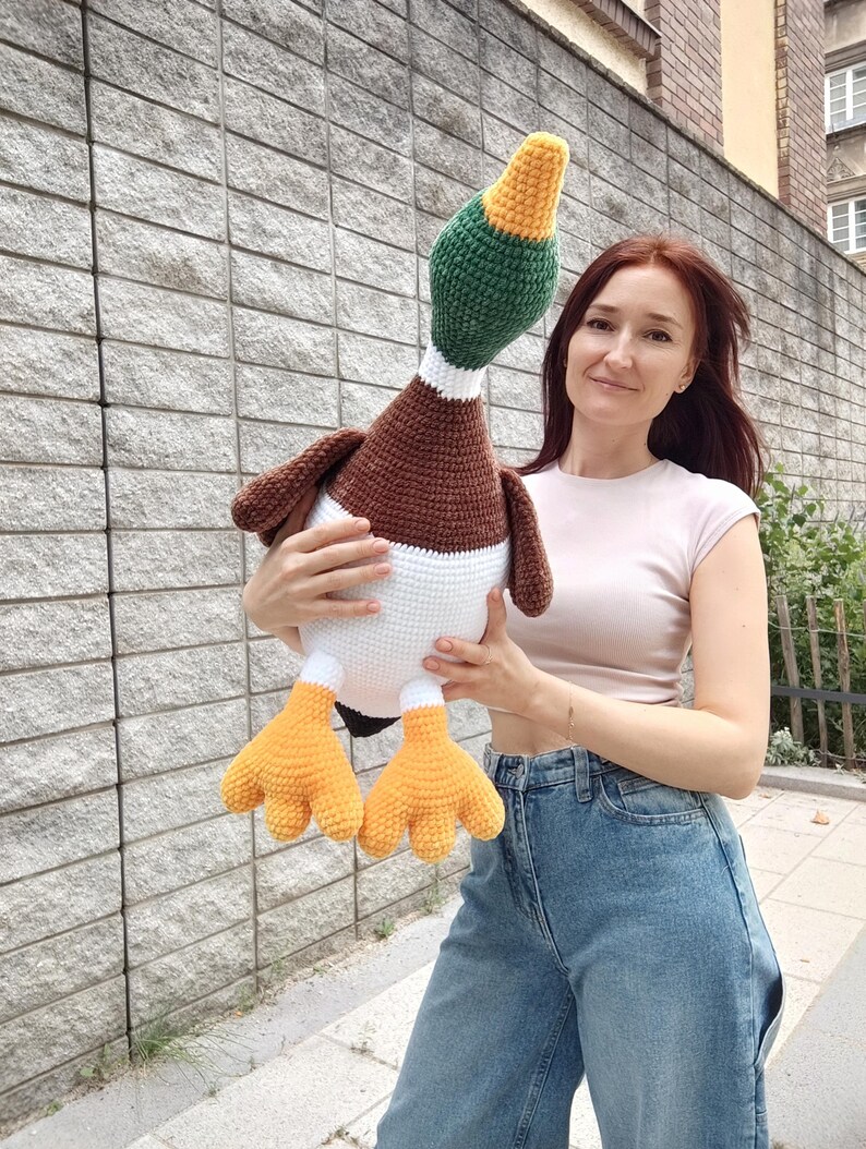 May include: A large, handmade crochet duck plushie. The duck features a brown body, white chest, green head, and yellow beak and feet. The plush toy is held in front of a gray stone wall.