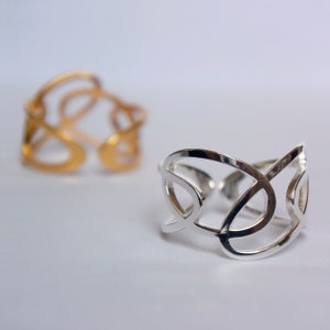 Può includere: Due anelli, uno in argento e uno in oro, con un designo traforato intricato. Gli anelli sono in metallo e hanno un motivo geometrico delicato.