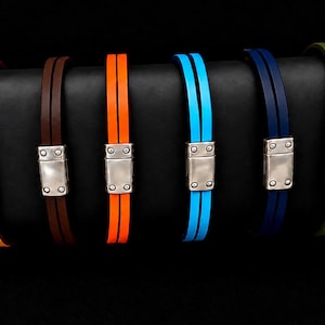 Könnte beinhalten: Eine Kollektion von acht doppelsträngigen Armbändern in verschiedenen Farben wie Orange, Braun, Blau, Grün und Schwarz. Jedes Armband verfügt über einen rechteckigen, silberfarbenen magnetischen Metallverschluss mit vier sichtbaren Nieten.