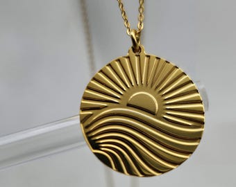 May include: A gold necklace with a round pendant featuring a sun rising over waves. The pendant has a textured design.