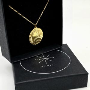 Peut inclure: Un collier en or avec un pendentif rond représentant un soleil et une vague. Le collier est dans une boîte noire avec le texte "OCTAGRAMME bijoux".