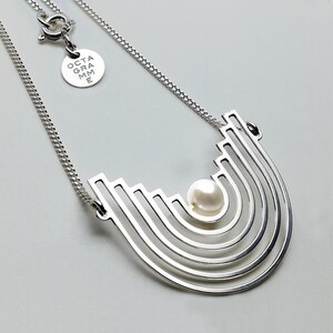 Peut inclure: Un collier en argent avec un pendentif géométrique orné d'une seule perle au centre. Le pendentif est en forme d'une série d'arches concentriques, créant un design moderne et abstrait.